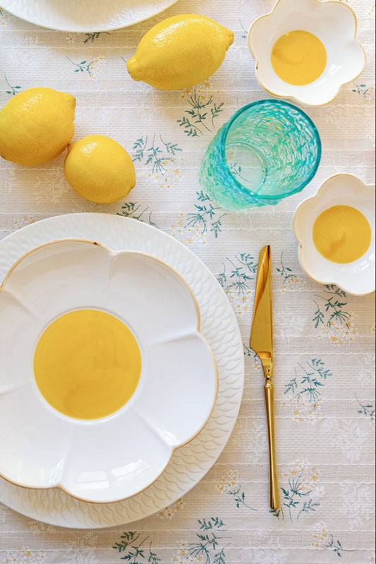 A set table with white flower-shaped plates, yellow centers, and blue glassware, surrounded by lemons and floral tablecloth.