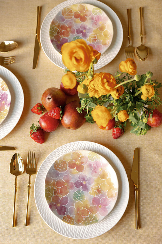 Decorative table setting with floral plates, fruits, and flowers on a beige tablecloth.