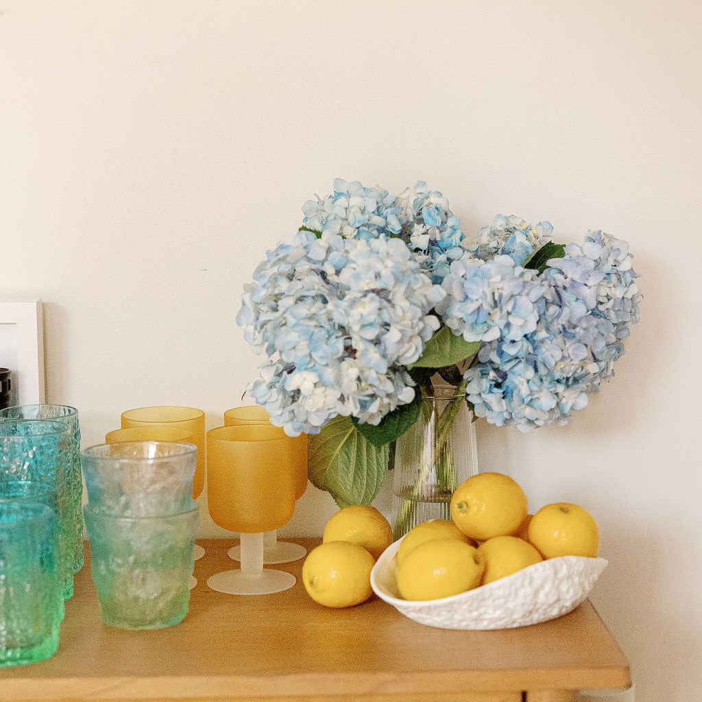 Frosted amber and blue Sea Glass cups on a wood cabinet styled with blue hydrangeas and lemons.