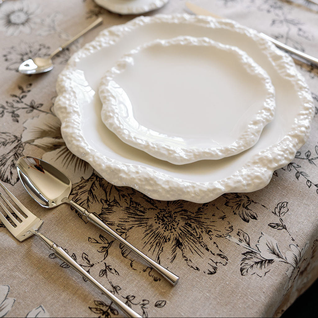 Polished silverware beside textured white scalloped plates on a floral toile tablecloth.