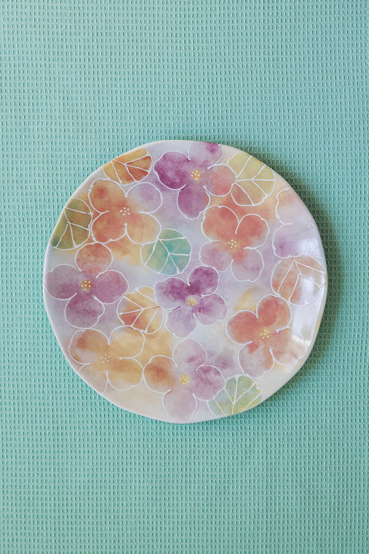 A round ceramic plate with a watercolor floral pattern in shades of purple and green on a seafoam green background
