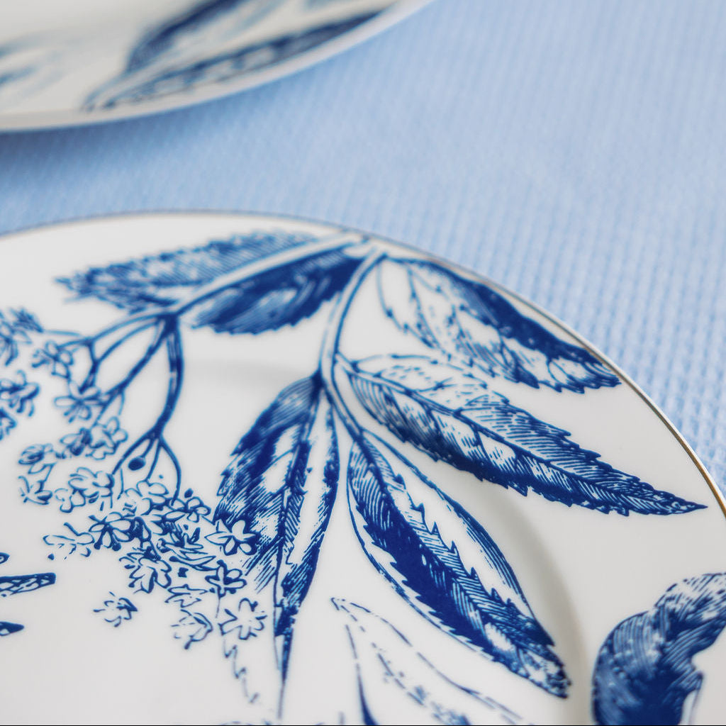 Close-up of two Tuscany gilded porcelain plates with blue toile leaf print and gold rims on a pale blue cloth.