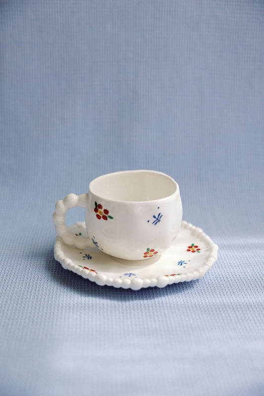 A white ceramic tea cup with blue and red floral patterns and a saucer with a scalloped edge