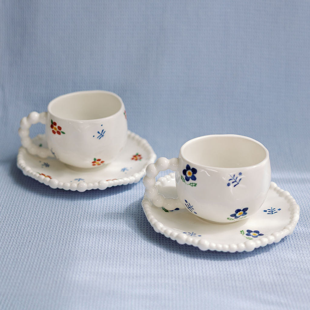 Pair of Cozy Blossom cups with blue and orange flowers and beaded-edge saucers on a blue cloth backdrop.