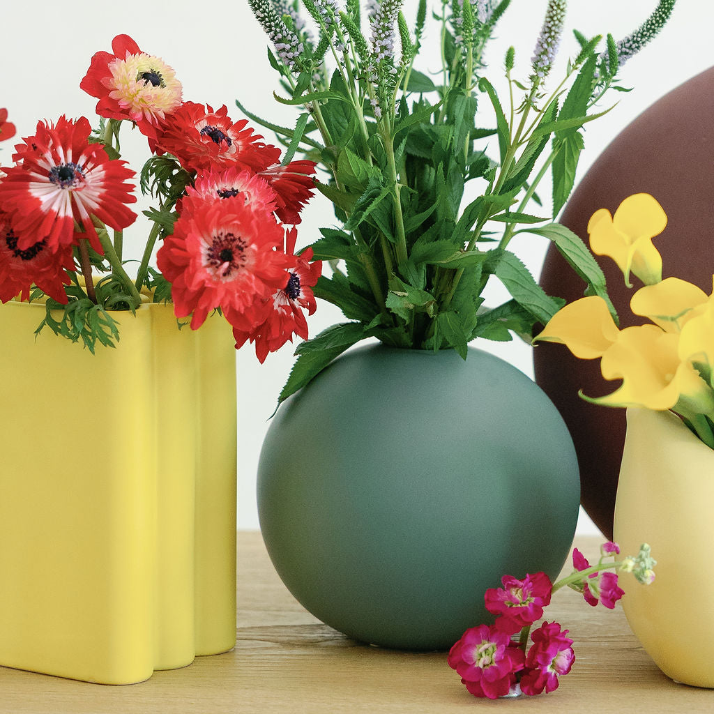 Yellow accordion vase with red anemones grouped beside green and brown round vases on a wood surface.
