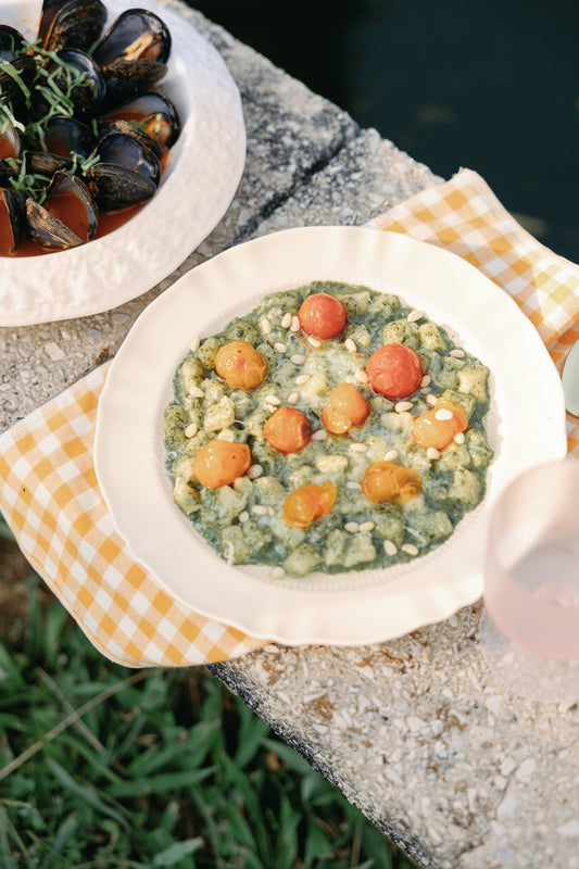 Provence Pleated scalloped white plate holding gnocchi with cherry tomatoes on a yellow gingham napkin outdoors.