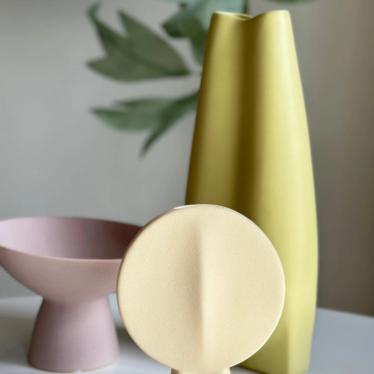 Bifold ceramic vase in cream with folded disc shape, styled beside a chartreuse vase and pink pedestal bowl