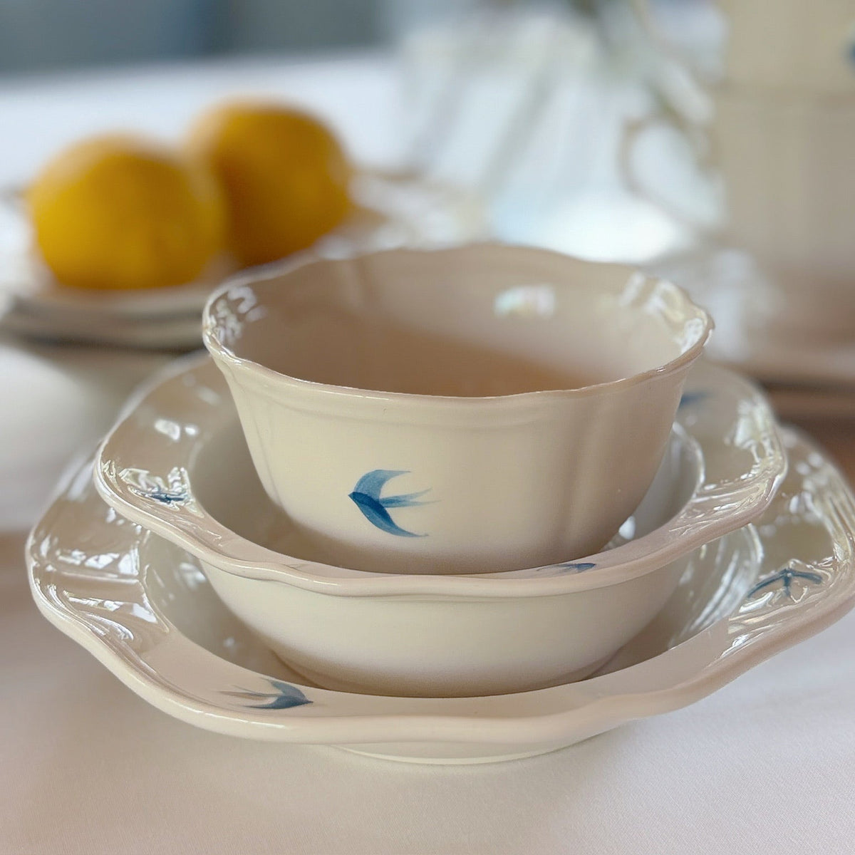 A stack of cream-colored ceramic tableware, including a small bowl with a blue bird motif, nested in a deeper bowl and a matching scalloped saucer, set on a table with a softly blurred background.