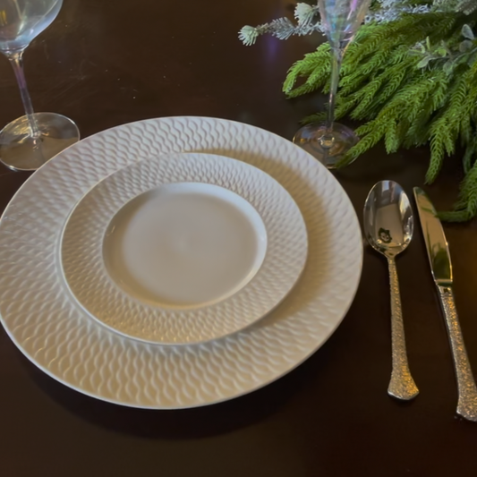 A close-up of a white ceramic dinner plate set with a textured, dimpled pattern, accompanied by silver-toned flatware on a dark wooden table with green foliage and wine glasses.