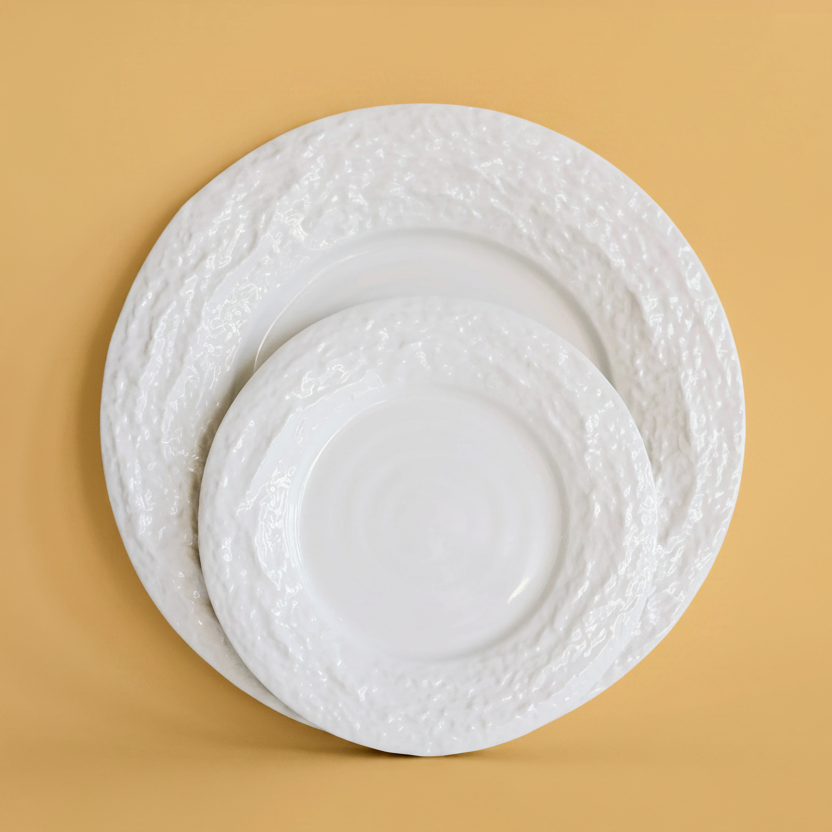 two white ceramic plates with textured, hammered-style rims stacked and overlapping on a solid mustard yellow background.