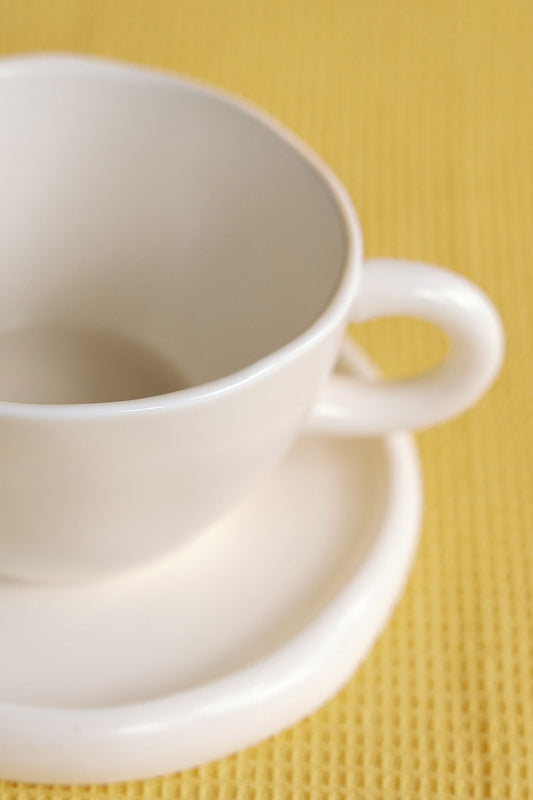 Bright Side matte white cup and saucer close-up showing the looped round handle on a yellow woven mat.