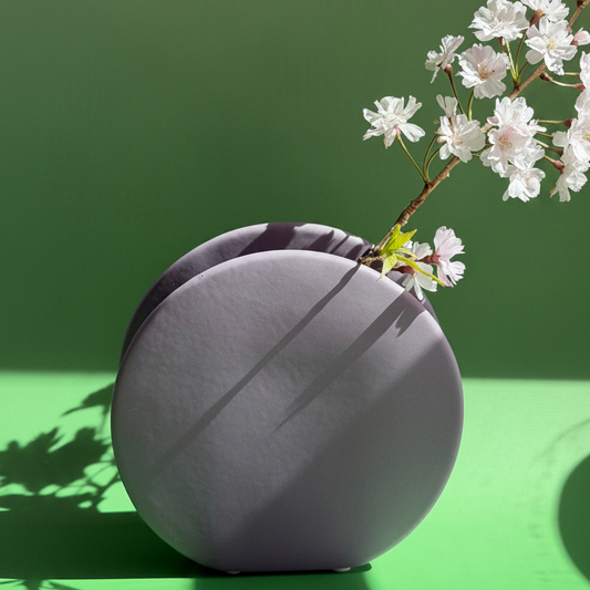 Ceramic Circle Bud Vase in grey, round flat vase holding white blossom sprigs against a green backdrop.