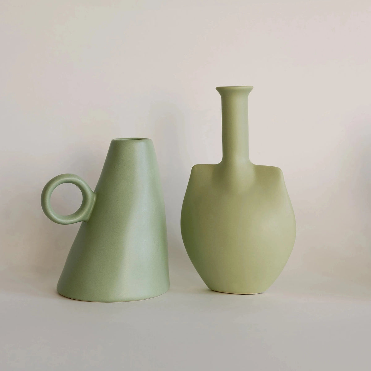 Sage green conical bud vase with loop handle beside a sage trowel-shaped bottle vase, two matte ceramic vases side by side