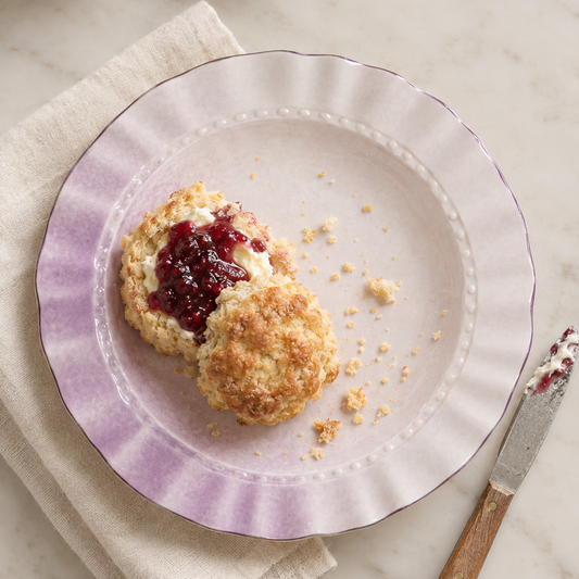 Two golden-brown scones topped with dark red berry jam on a lavender-rimmed plate, set on a cream napkin with a knife and bowls of jam and cream in the background.