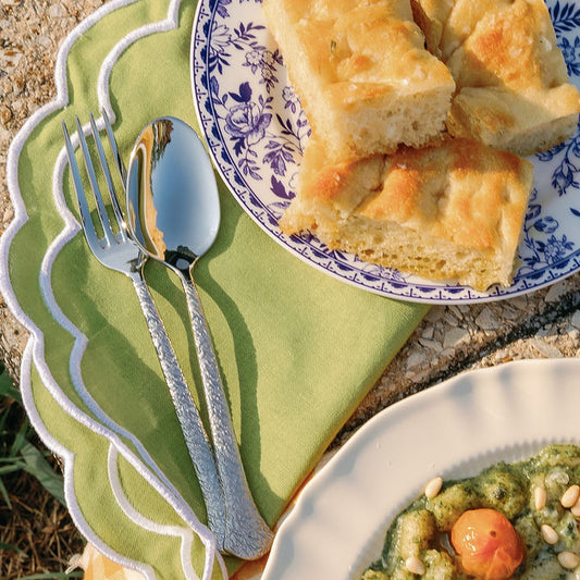 Hammered stainless steel fork and spoon styled on a green napkin beside a blue and white plate of focaccia outdoors.