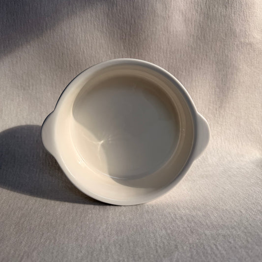 Overhead view of a small white ribbed baking bowl with side handles on soft fabric.
