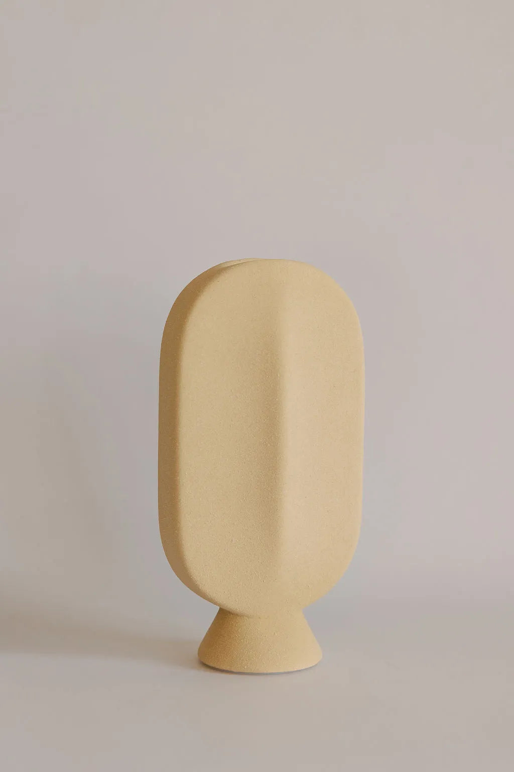 Beige ceramic bud vase in flattened oval shape on a small pedestal foot, matte sandy finish