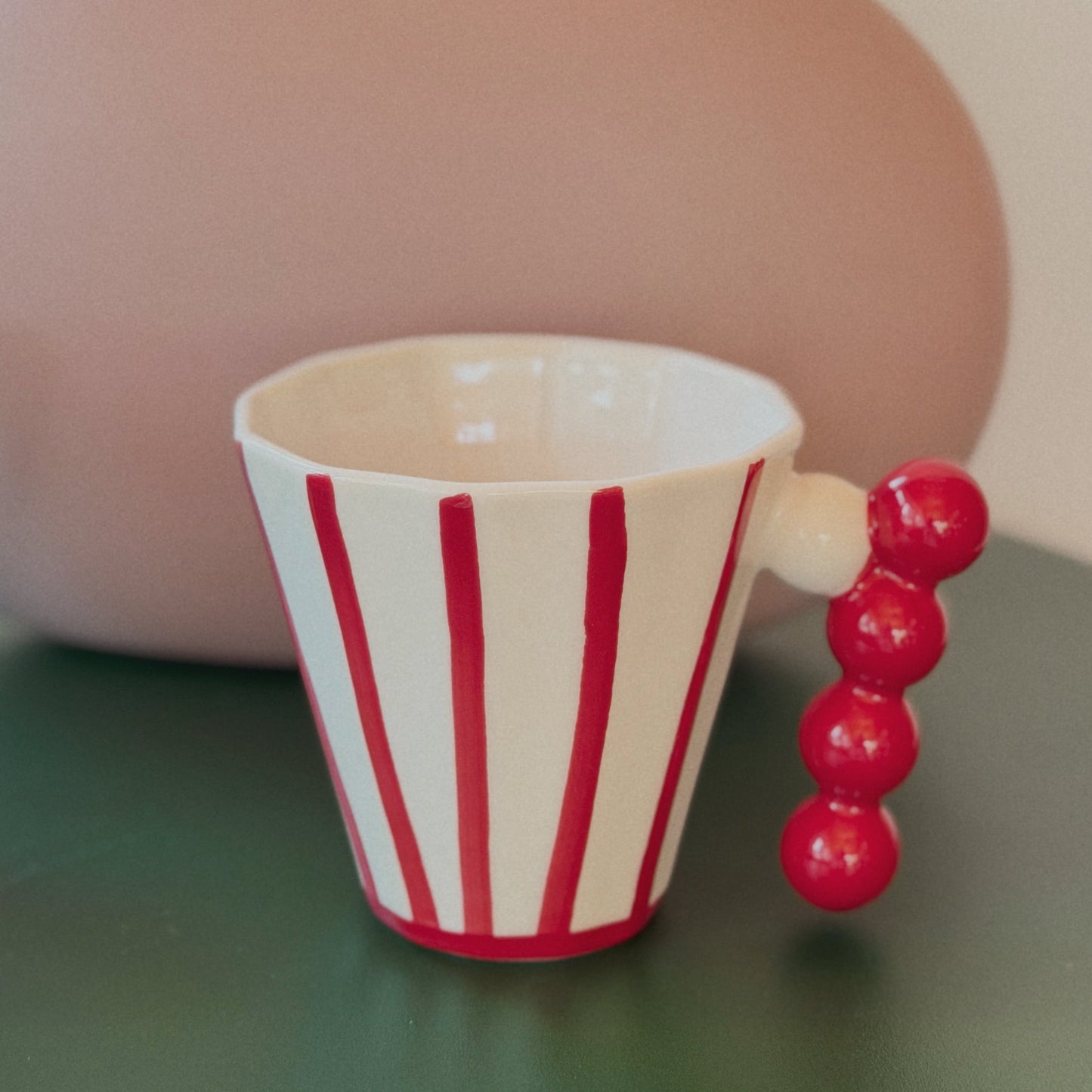 Striped cup with a beaded handle on a green surface, blurred pink vase in the background