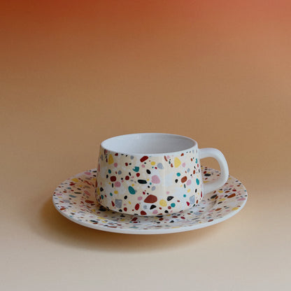 Terrazzo tea cup and saucer with colorful confetti speckles against a warm orange gradient backdrop.
