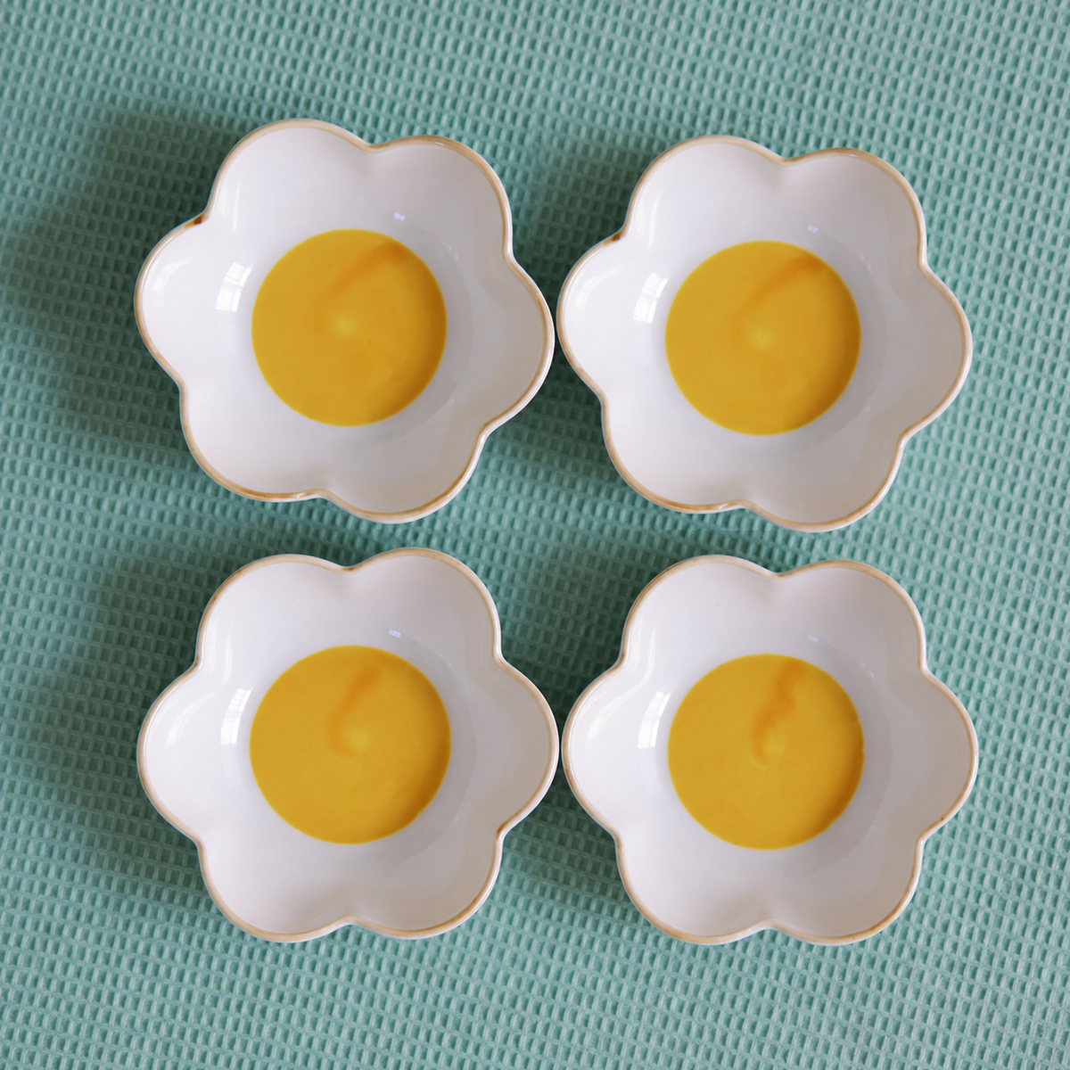 Four small white flower-shaped ceramic bowls with gold trim, arranged in a 2x2 grid on a textured mint green surface. Each bowl is filled with a yellow-orange puree or cream.