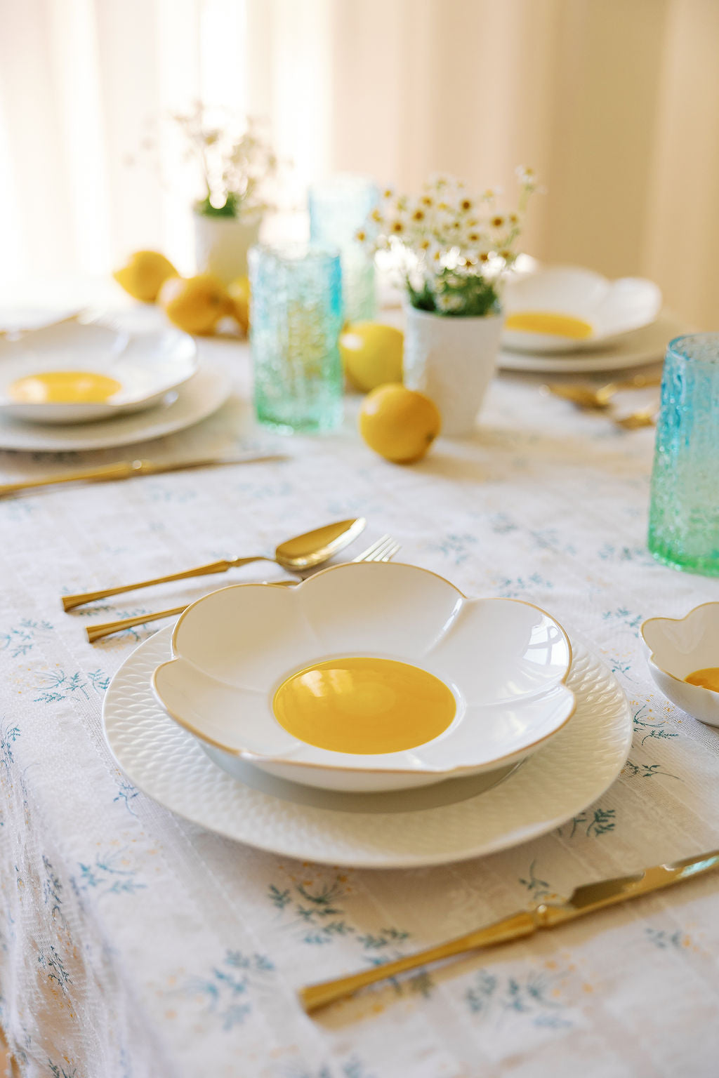 Gold silverware at a place setting with daisy-shaped plates, blue glassware and lemons on floral linen.