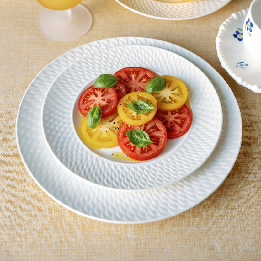 White textured ceramic dinnerware set on a beige tablecloth, featuring a large charger plate, a dinner plate, and a bowl holding sliced red and yellow tomatoes with fresh basil leaves. A yellow goblet, lemons, and a blue hydrangea are arranged around it.