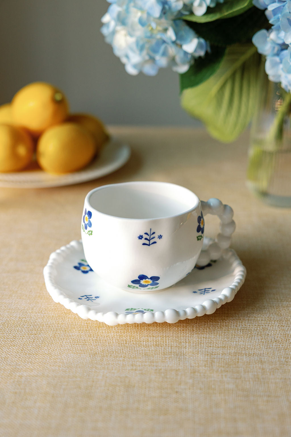 A white ceramic tea cup with blue floral patterns and a saucer with a scalloped edge, displayed on a table with a vase of flowers and lemons in the background.