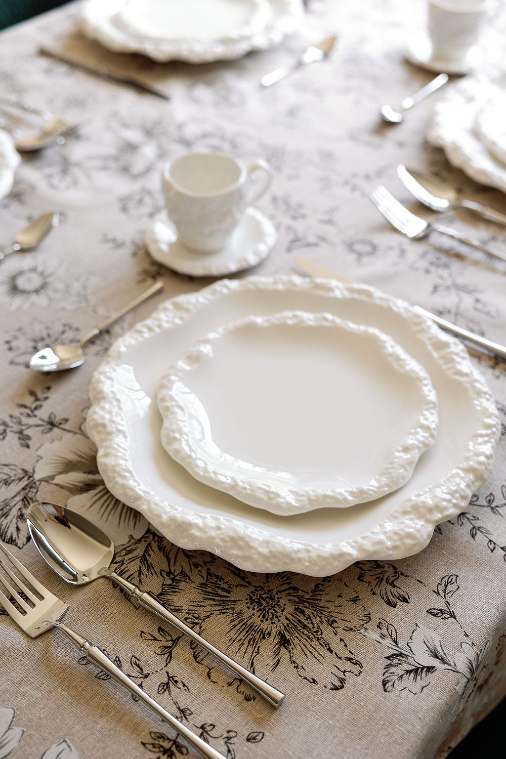 Polished silverware beside textured white scalloped plates on a floral toile tablecloth.
