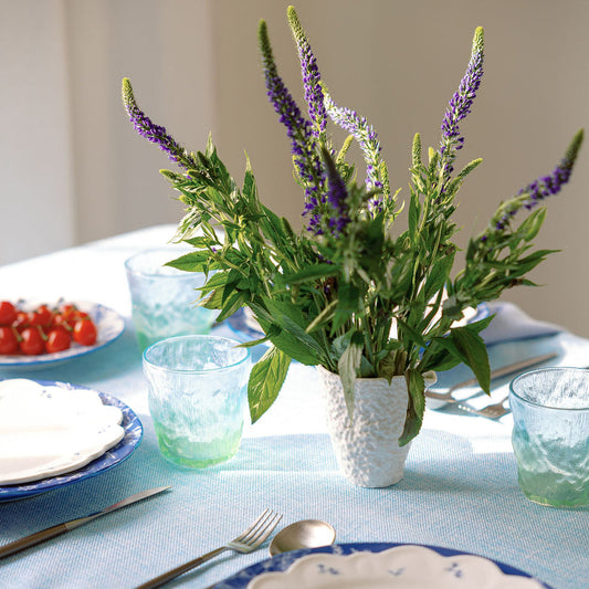 A styled dining table setting with a central bouquet of green leaves and purple flowers in a textured white vase, surrounded by blue and white plates, light blue textured glasses, silver cutlery, and a light blue tablecloth.