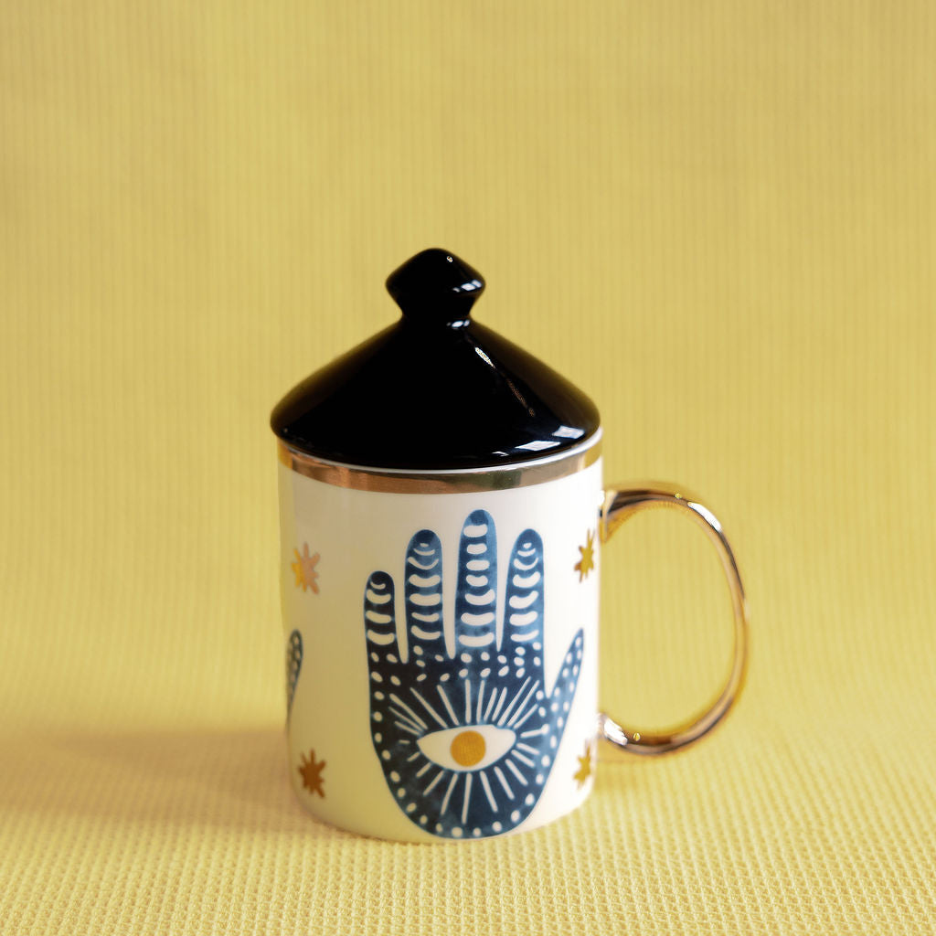 Witching Hour mug painted with a blue hamsa hand and evil eye, gold handle and black lid on yellow.
