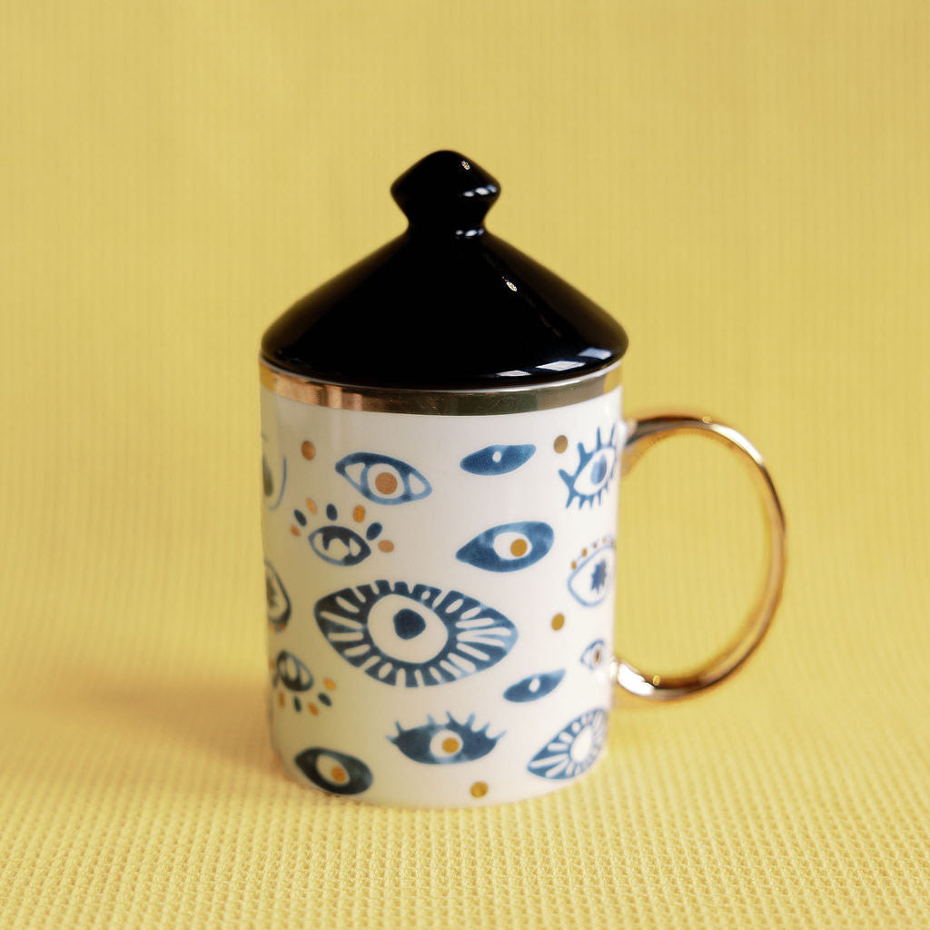 Witching Hour porcelain mug with blue evil-eye pattern, gold handle and black peaked lid on a yellow backdrop.
