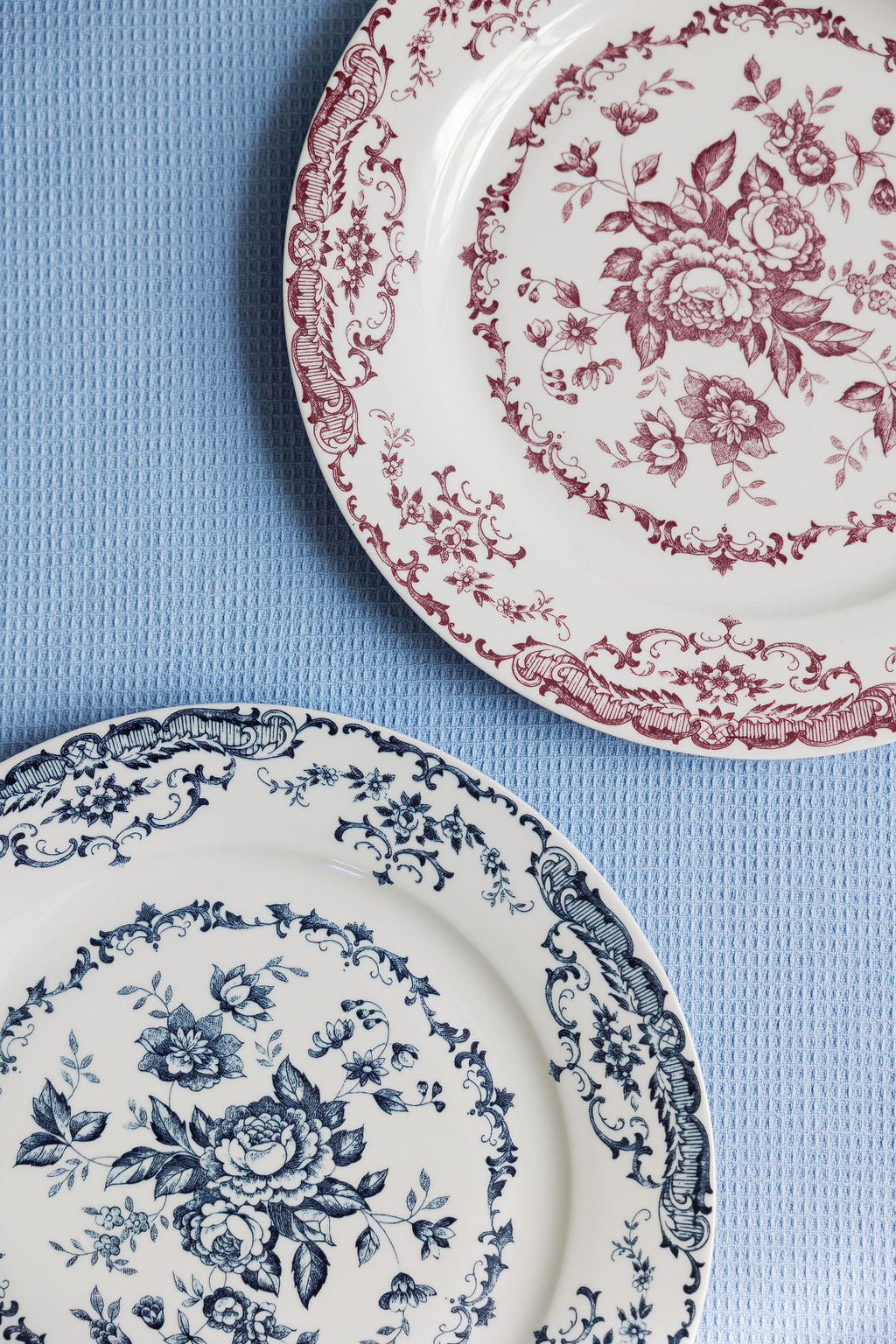 Two Chantilly Lace porcelain plates, one red floral and one blue floral, side by side on a pale blue surface.