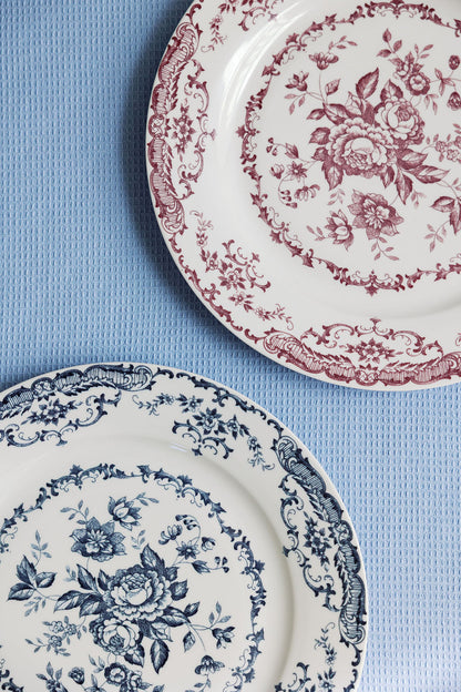 Two Chantilly Lace porcelain plates, one red floral and one blue floral, side by side on a pale blue surface.