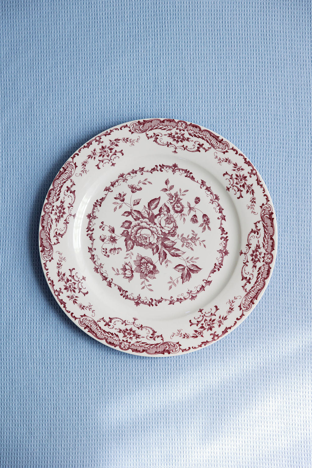 Chantilly Lace porcelain dinner plate with red floral and scrollwork border on a pale blue textured backdrop.