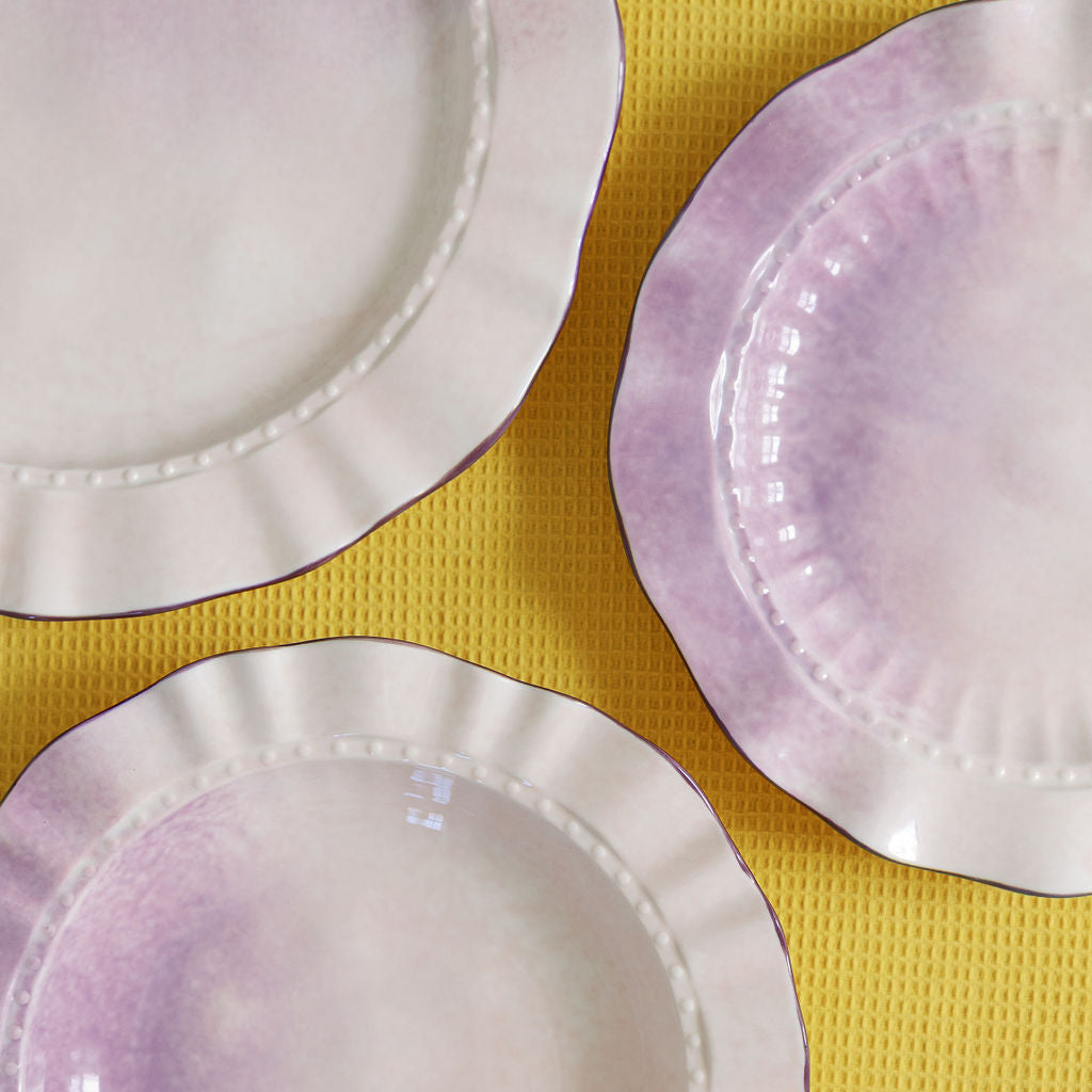 Three scalloped ceramic plates with a white and light purple watercolor wash, arranged on a textured yellow background.