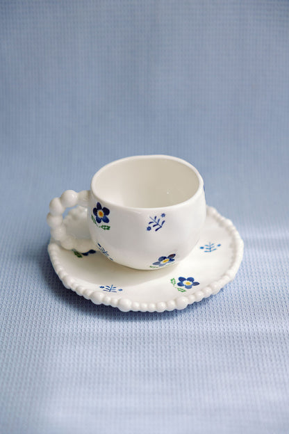 A white ceramic tea cup with blue floral patterns and a saucer with a scalloped edge.