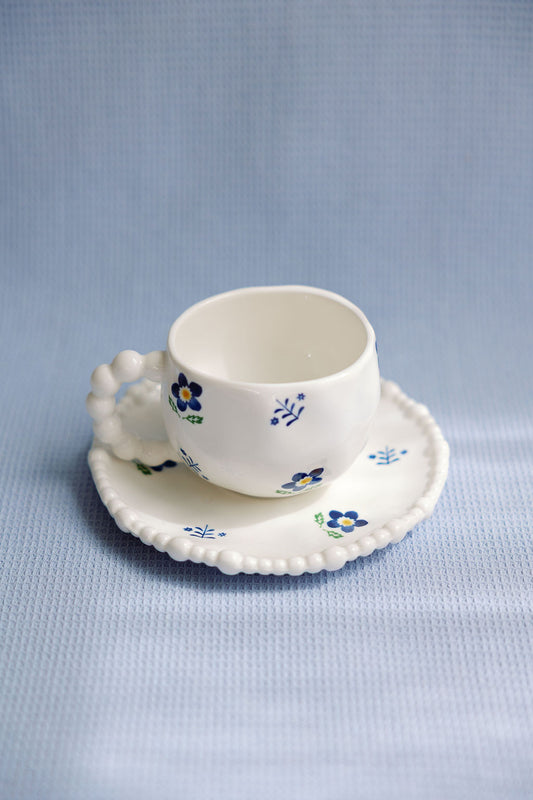A white ceramic tea cup with blue floral patterns and a saucer with a scalloped edge.