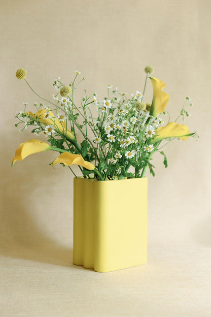 Butter yellow accordion ceramic vase holding calla lilies and daisies against a linen backdrop.
