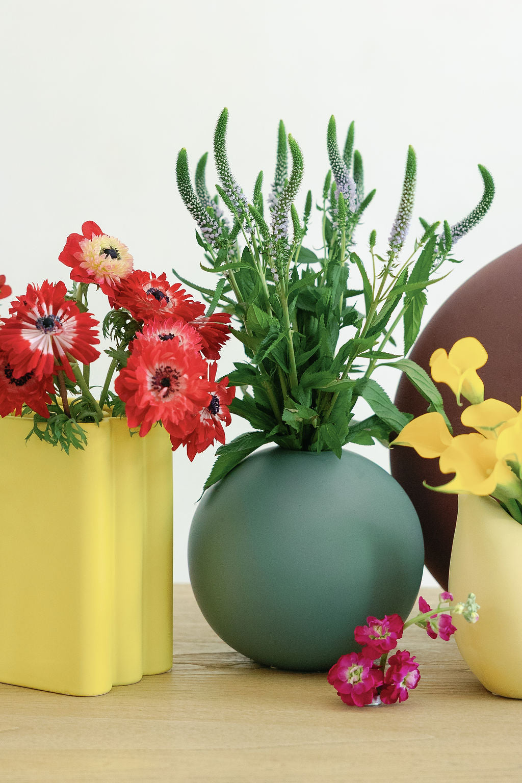 Yellow accordion vase with red anemones grouped beside green and brown round vases on a wood surface.