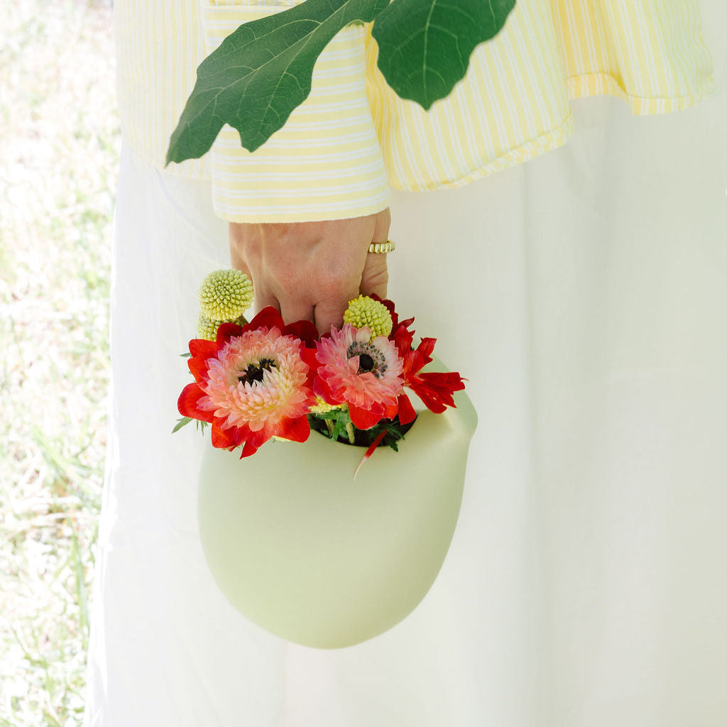 A person in a pale yellow and white striped dress holds a mint green basket-shaped vase filled with red and pink flowers, with a large green leaf hanging over the scene.