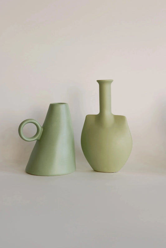 Sage green conical bud vase with loop handle beside a sage trowel-shaped bottle vase, two matte ceramic vases side by side