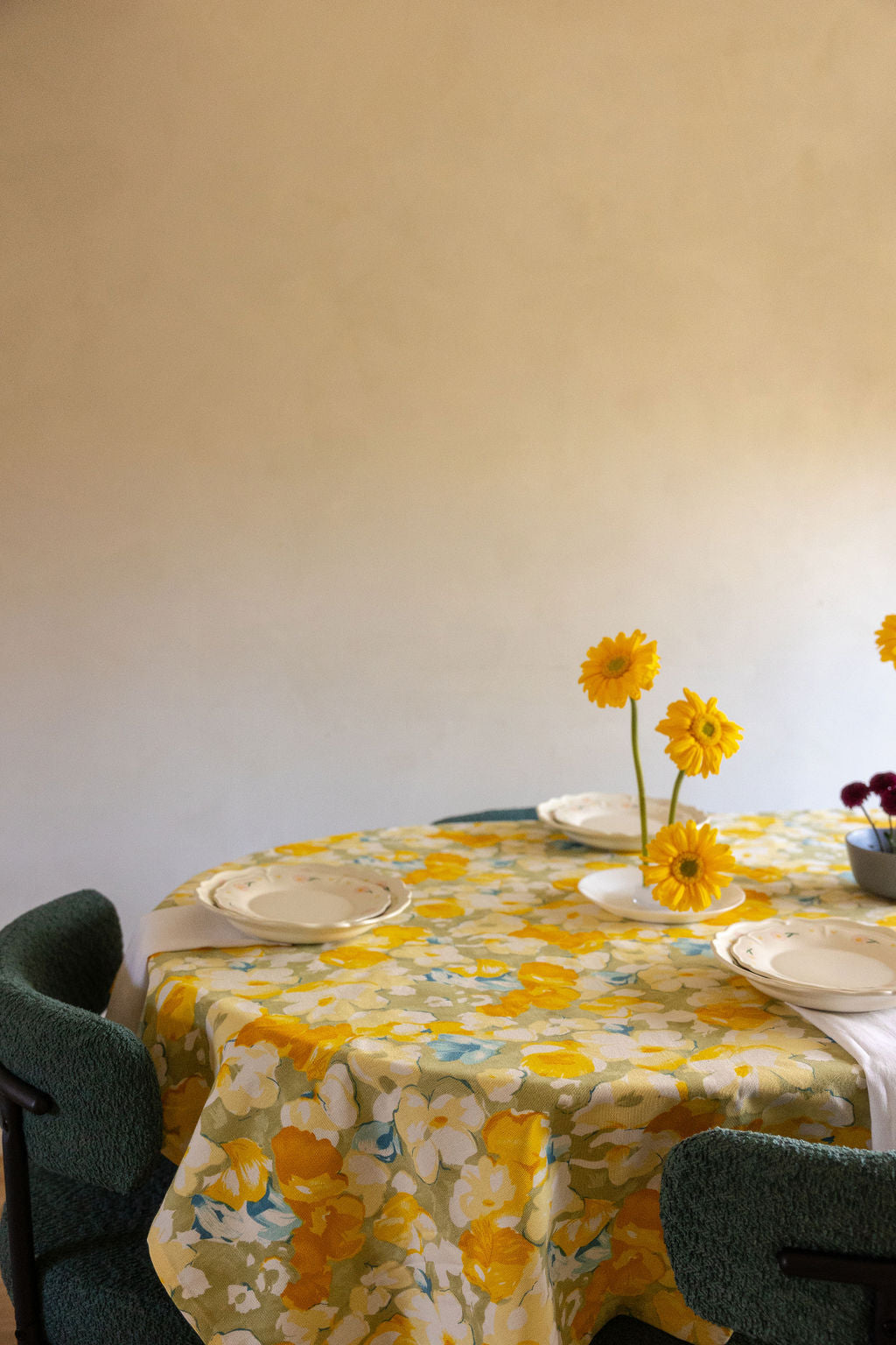 Round table set with Coquette Picnic floral plates, yellow daisies in a vase and green boucle chairs.