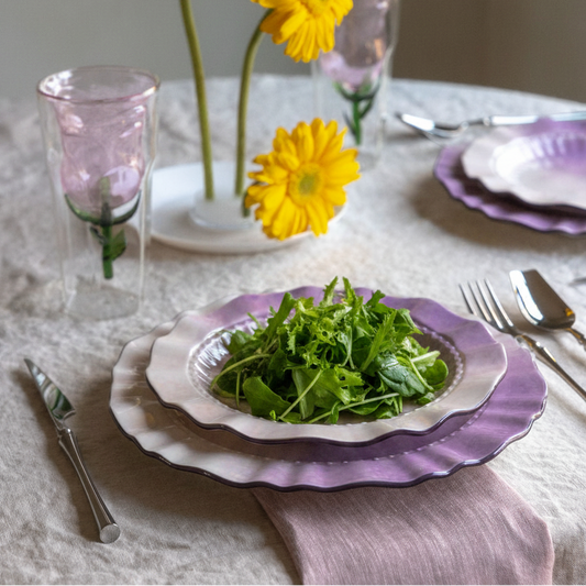 A styled table setting with lavender and white plates holding a green salad, pink glass tumblers, silver cutlery, and three yellow flowers in a vase against a soft neutral background.