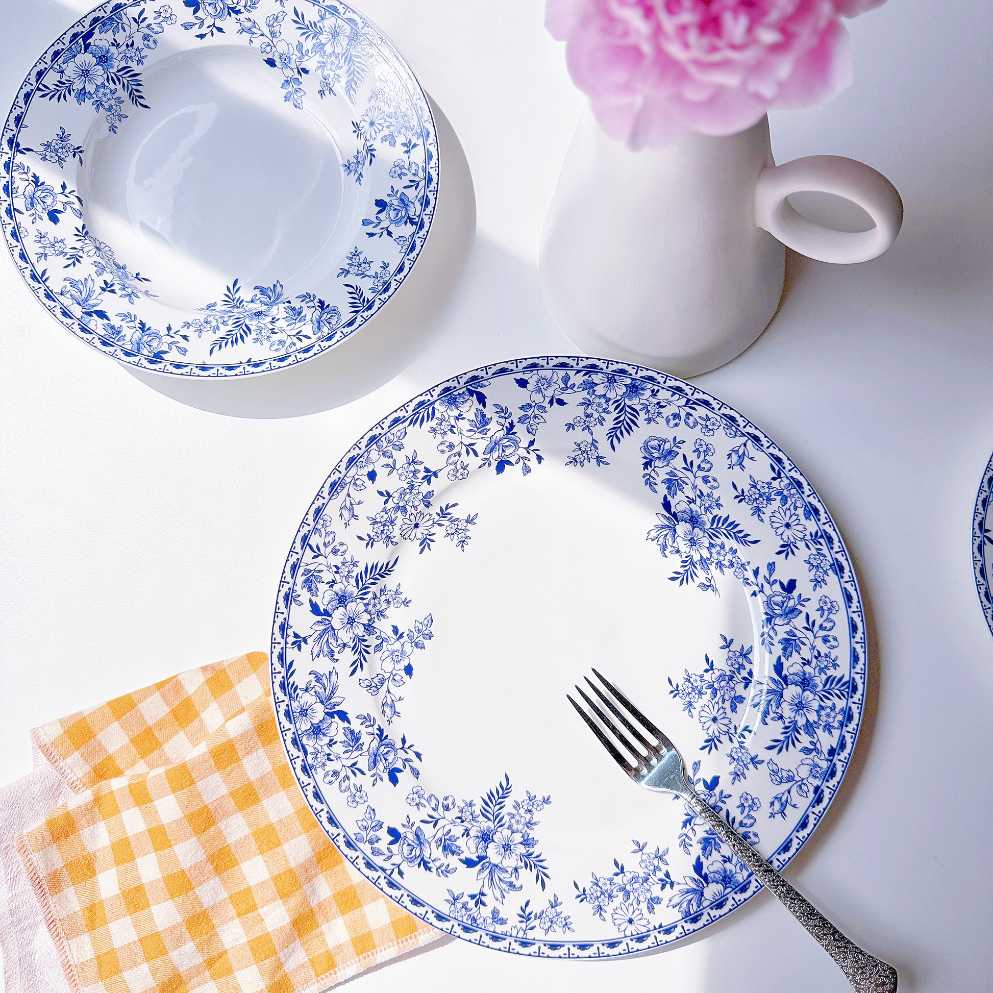 Set of blue floral-patterned plates with a fork, checkered napkin, and pink flower on a white background