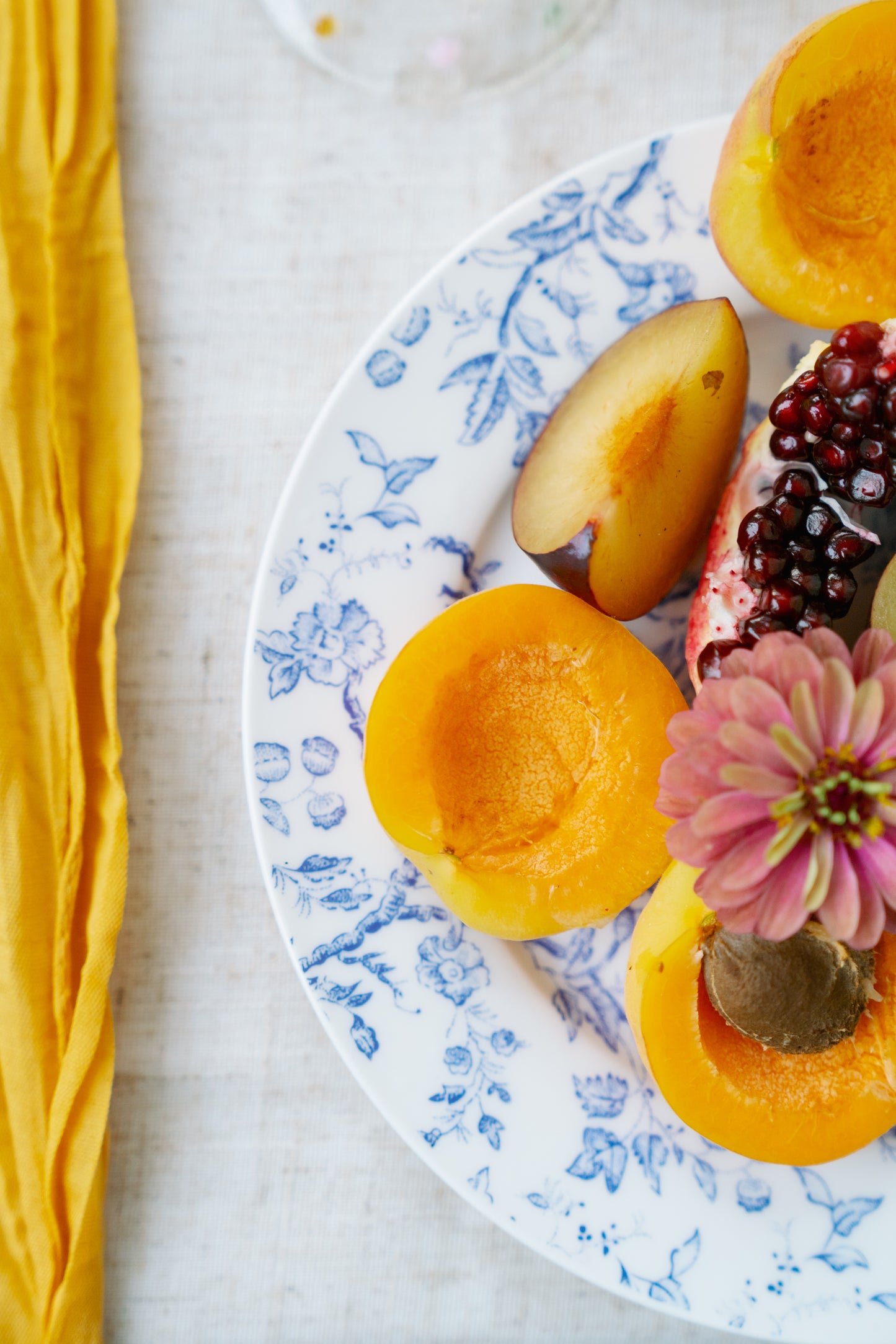Cottage Garden blue floral plate styled with sliced apricots, plum and pomegranate beside a yellow napkin.