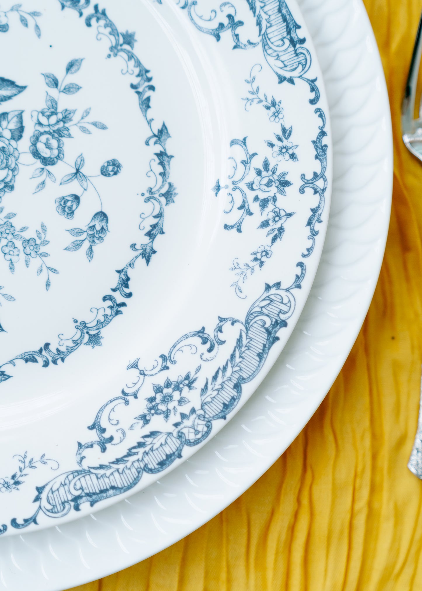 Close-up of blue floral and scroll detail on a Chantilly Lace plate over a ribbed white charger on yellow wood.