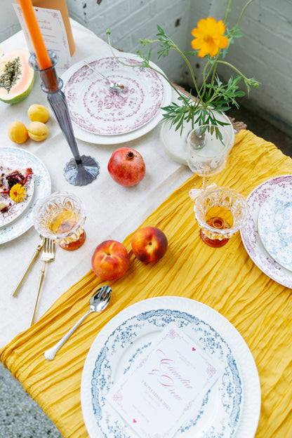 Chantilly Lace plates in red and blue florals set on a mustard runner with fruit and amber glasses, table scene.