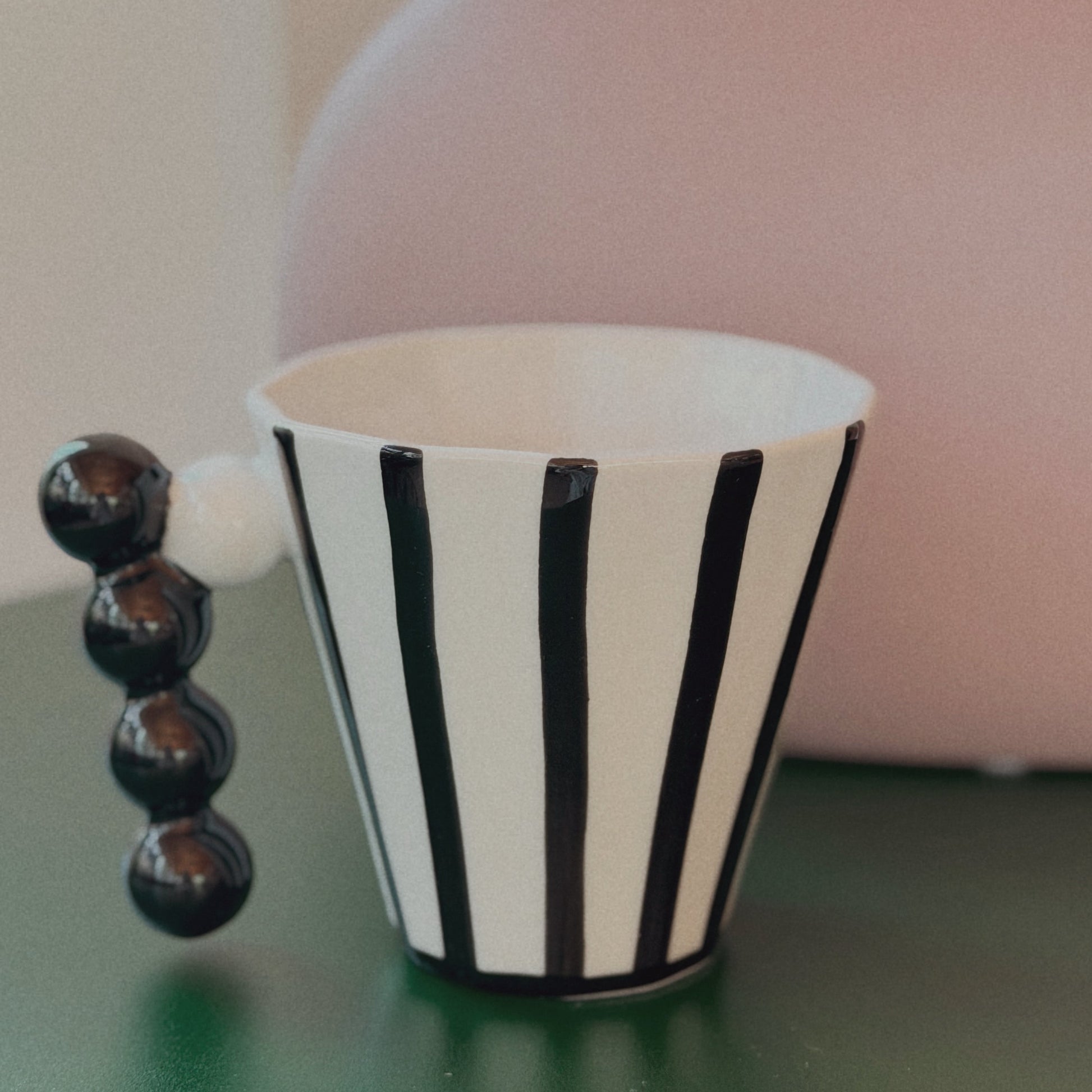 Black and white striped cup with a beaded handle on a green surface, blurred pink vase in the background