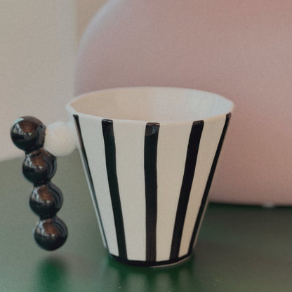 Black and white striped cup with a beaded handle on a green surface, blurred pink vase in the background