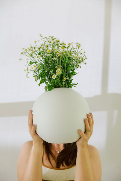 Person holding a large white vase filled with flowers against a light background